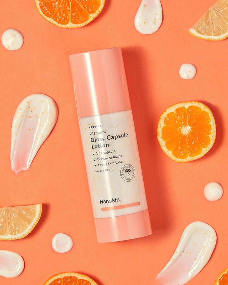 Hanskin Vitamin C Glow Capsule Lotion - lifestyle image of the lotion bottle on an orange background surrounded by slices of orange fruit and swipes of the lotion to showcase texture